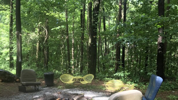 A forested campsite with a fire pit, chairs, and a table.