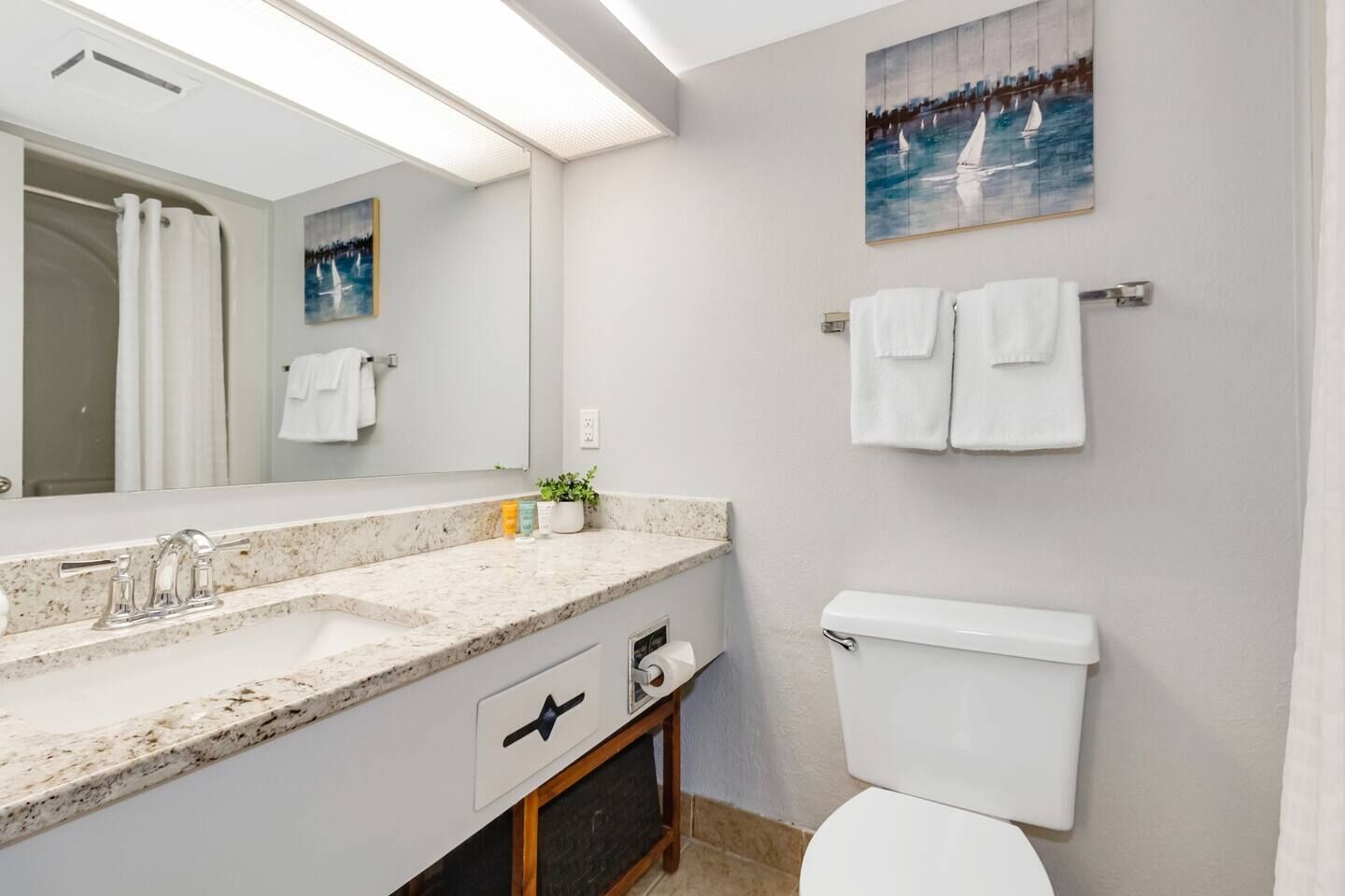 A modern bathroom with a marble sink, a toilet, and a shower. There are towels hanging on the wall and a painting above the toilet.