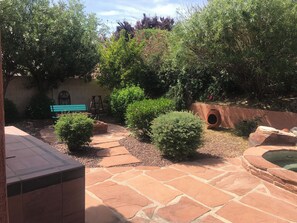 Un patio con suelo de piedra, un banco verde y varios arbustos.
