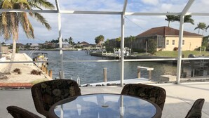 A covered outdoor seating area with wicker chairs and a glass table, overlooking a marina with boats and a building in the background.