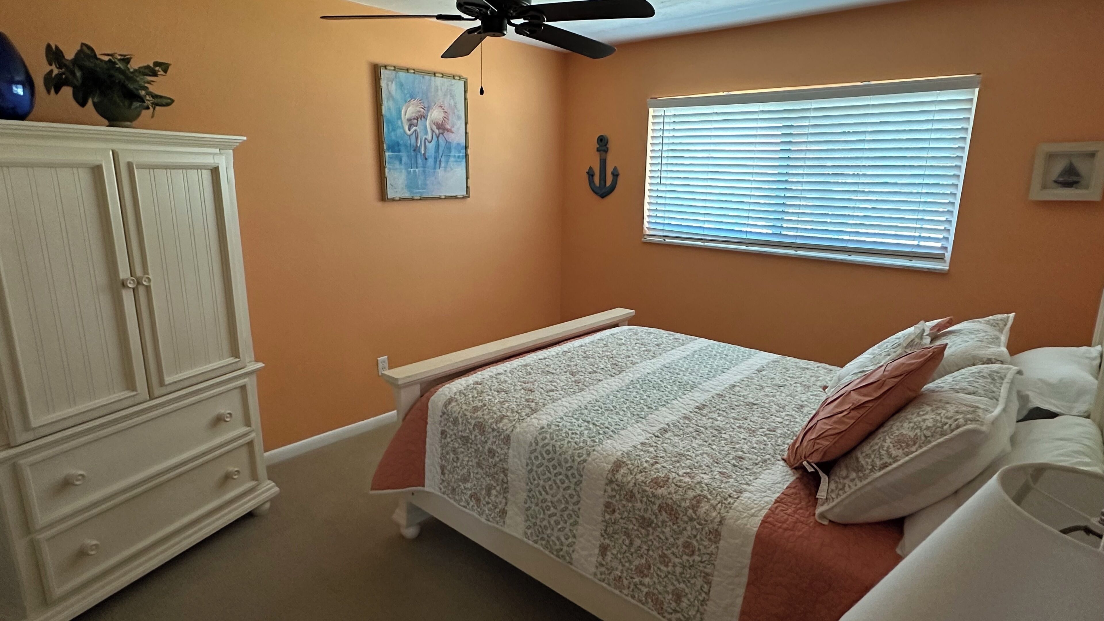 A bedroom with a bed, a ceiling fan, a window with blinds, a painting, and a cabinet.