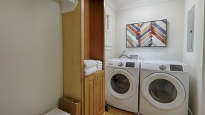 Une buanderie équipée de deux machines à laver à chargement frontal, d’un meuble en bois contenant des serviettes et d’un tableau abstrait aux couleurs vives accroché au mur.