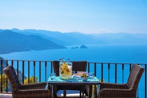 Ein Balkon mit Korbstühlen und einem Tisch für zwei Personen, mit Blick auf die Küste, Berge und das Meer.