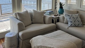 Tunnelmallinen olohuone, jossa on suuri ikkuna, josta avautuu näkymä laiturille. Huoneessa on beige kulmasohva, pieni sivupöytä, jossa on lamppu ja koriste-esineitä, sekä kudottu matto.