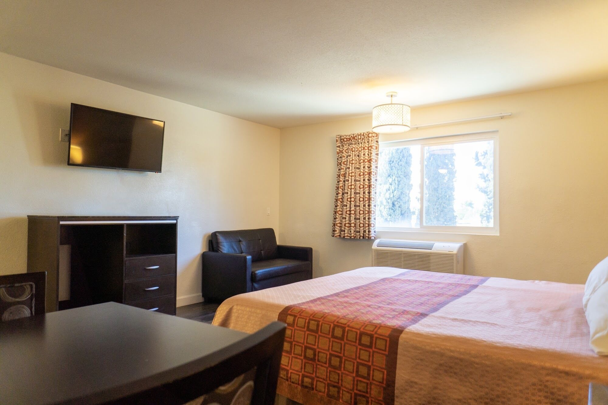 Quarto de hotel com cama, escrivaninha, cadeira, televisão e janela com cortinas.