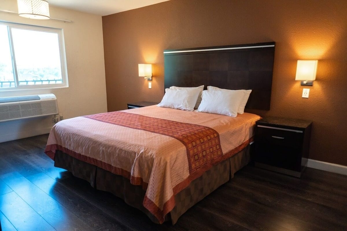 A hotel room with a large bed, two bedside tables, a nightstand, a window, and an air conditioning unit.
