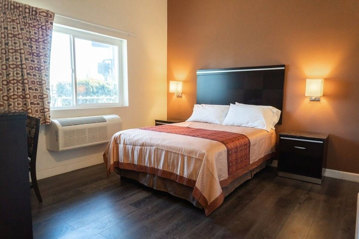 A hotel room with a bed, two bedside tables, a window with curtains, and an air conditioning unit.
