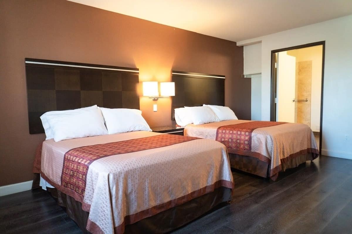 A hotel room with two beds, each with a patterned bedspread and white pillows. There are wall-mounted lights above the beds and a door leading to another room.