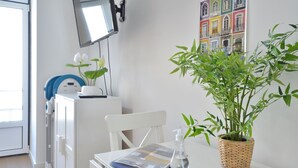 Ein Zimmer mit weißem Schreibtisch, einem Sessel, einem an der Wand befestigten Fernseher, einem Blumentopf und einem farbenfrohen Kunstwerk an der Wand.