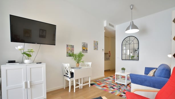 Ein modernes Wohnzimmer mit Flachbildfernseher, weißen Möbeln, einem farbenfrohen Teppich und einem blauen Sofa.