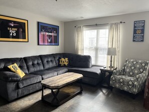 Ein Wohnzimmer mit einer Eckcouch, einem Couchtisch, einem gemusterten Sessel und gerahmten Sportmemorabilien an den Wänden.