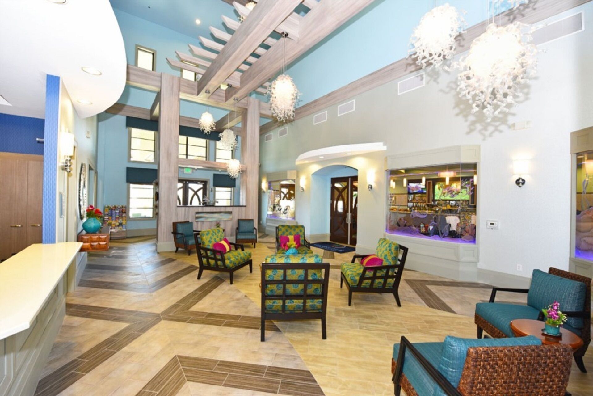 A spacious lobby with a high ceiling, a central seating area with colorful chairs, a bar area with a TV, and a large window providing natural light.