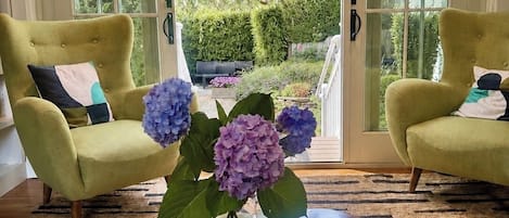 A room with a large wooden coffee table, two armchairs, and a view through glass doors to a garden.