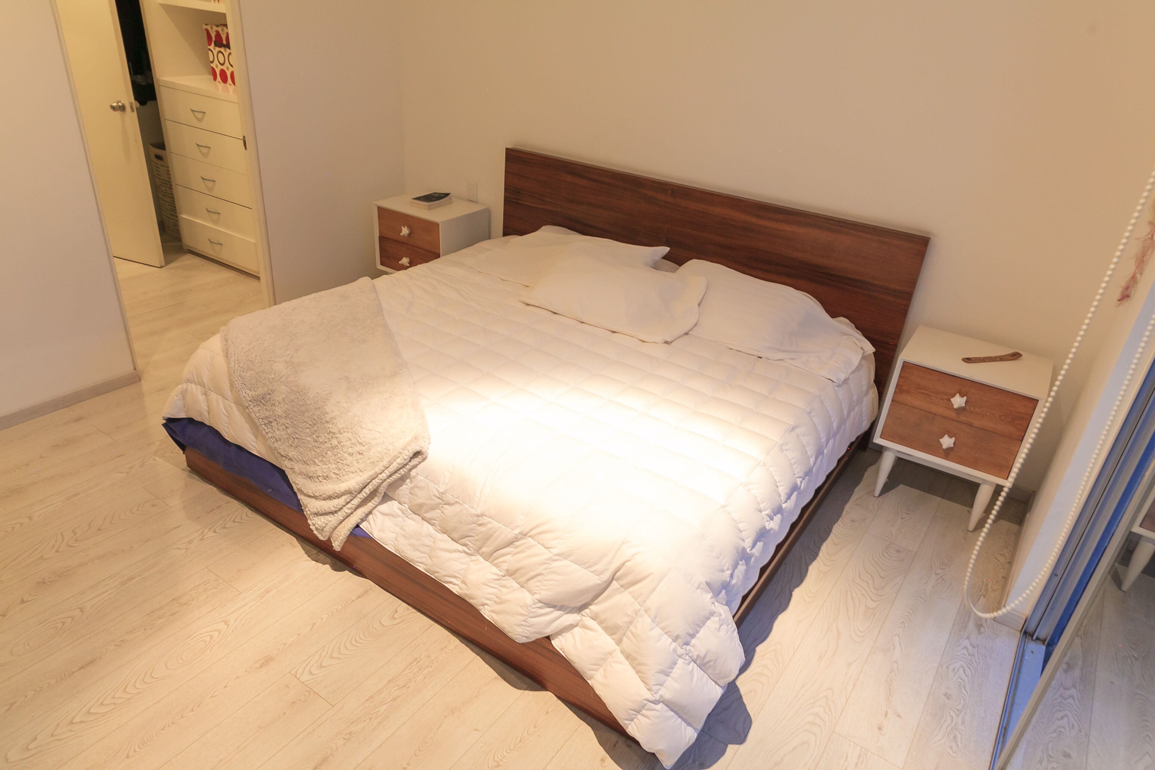 Een net opgemaakt bed met een houten hoofdbord, een nachtkastje met een telefoon, en een ladekast.