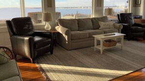 A living room with a sofa, chairs, a coffee table, and large windows offering a view of the sea and buildings. - HAMPTON BEACH OCEANFRONT HOME SITUATED ON GREAT BOARS HEAD (Hampton)