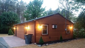 Una cabaña de madera con una puerta y dos ventanas, rodeada de árboles y un camino de grava.