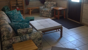Una sala de estar con sofá, dos sillones, una mesa de centro, una pequeña mesa de madera, un ventilador de techo, una ventana con cortinas y una puerta.