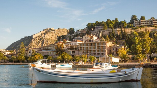 Un poble costaner amb un edifici destacat al penya-segat, barques amarrades al passeig marítim i un cel serè.