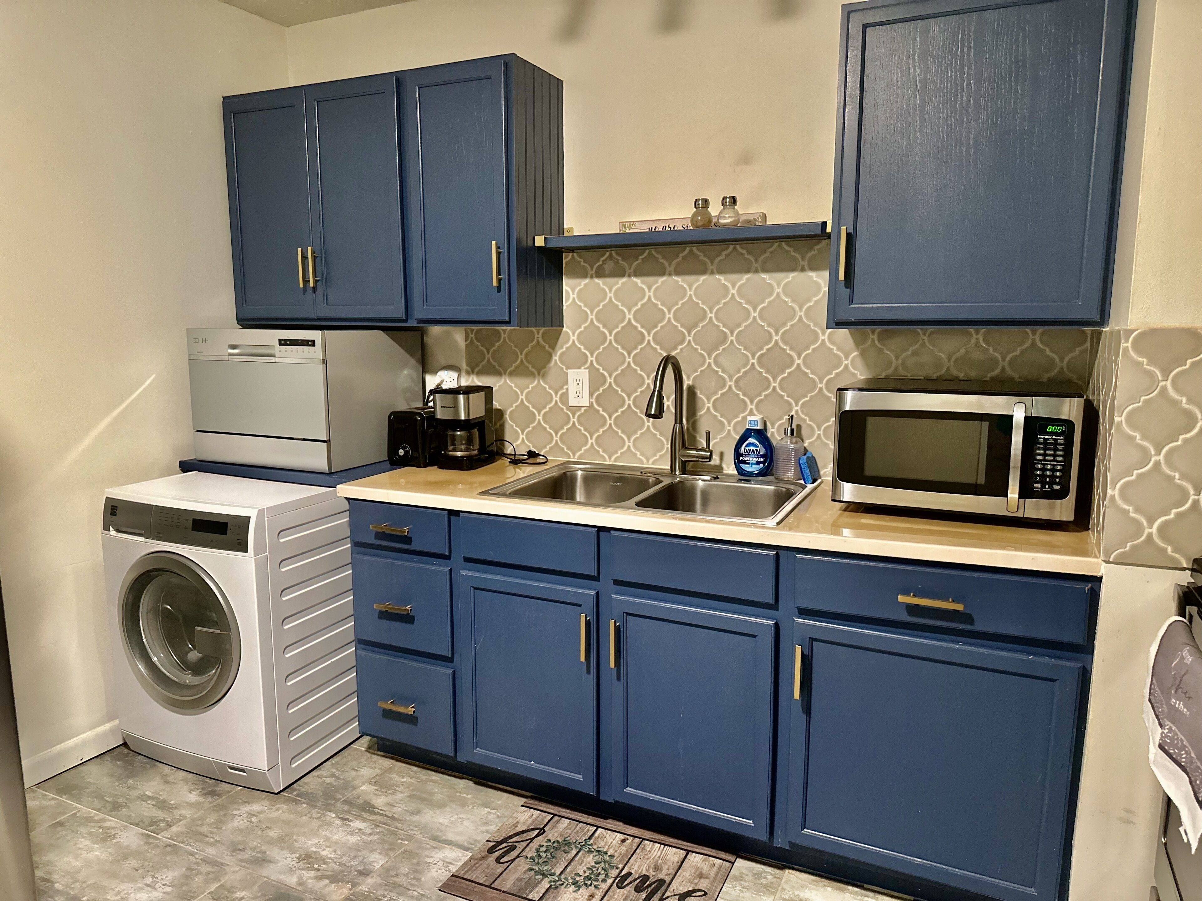 A compact kitchen with blue cabinets, a washing machine, a coffee maker, a microwave, and a sink.