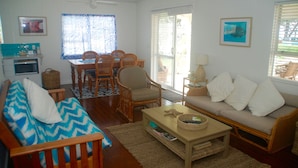 Uma sala de estar com sofá, mesa de centro, mesa de jantar com cadeiras, ventilador de teto e janela com persianas.