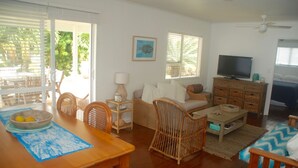 Uma sala de estar com uma mesa de jantar de madeira, cadeiras de vime, um sofá, uma TV e um ventilador de teto.