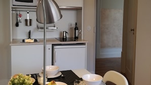 Een kleine keuken met een magnetron, koffiezetapparaat en een fles wijn. Op de voorgrond staat een eettafel met een schaal fruit en een kaars.