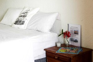 Una cama bien tendida con sábanas blancas y una almohada decorativa, una mesita de noche con una vela, un vaso de agua y un folleto turístico.