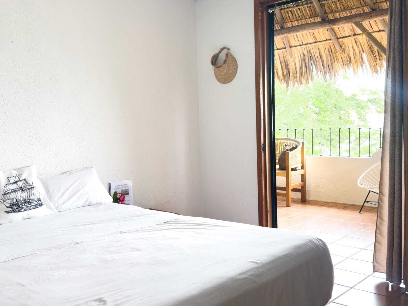 A bedroom with a bed, pillows, a small table with a book, and a view of an outdoor balcony with a chair and a thatched roof.