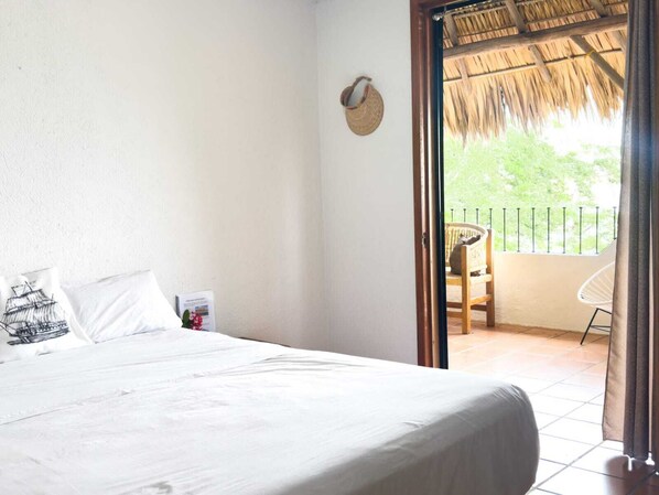 Un dormitorio con cama, almohadas, una mesita con un libro y vistas a un balcón exterior con una silla y techo de paja.