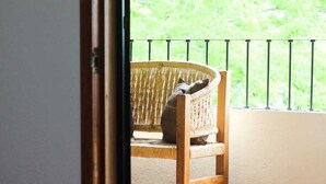 A room with a bed, a wicker chair, and a view of greenery through a window with a metal railing. - Acogedora palapa privada cerca de la playa. (Puerto Escondido)