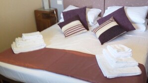 A neatly made bed with white linens, a brown bedspread, and several pillows in shades of purple and white.