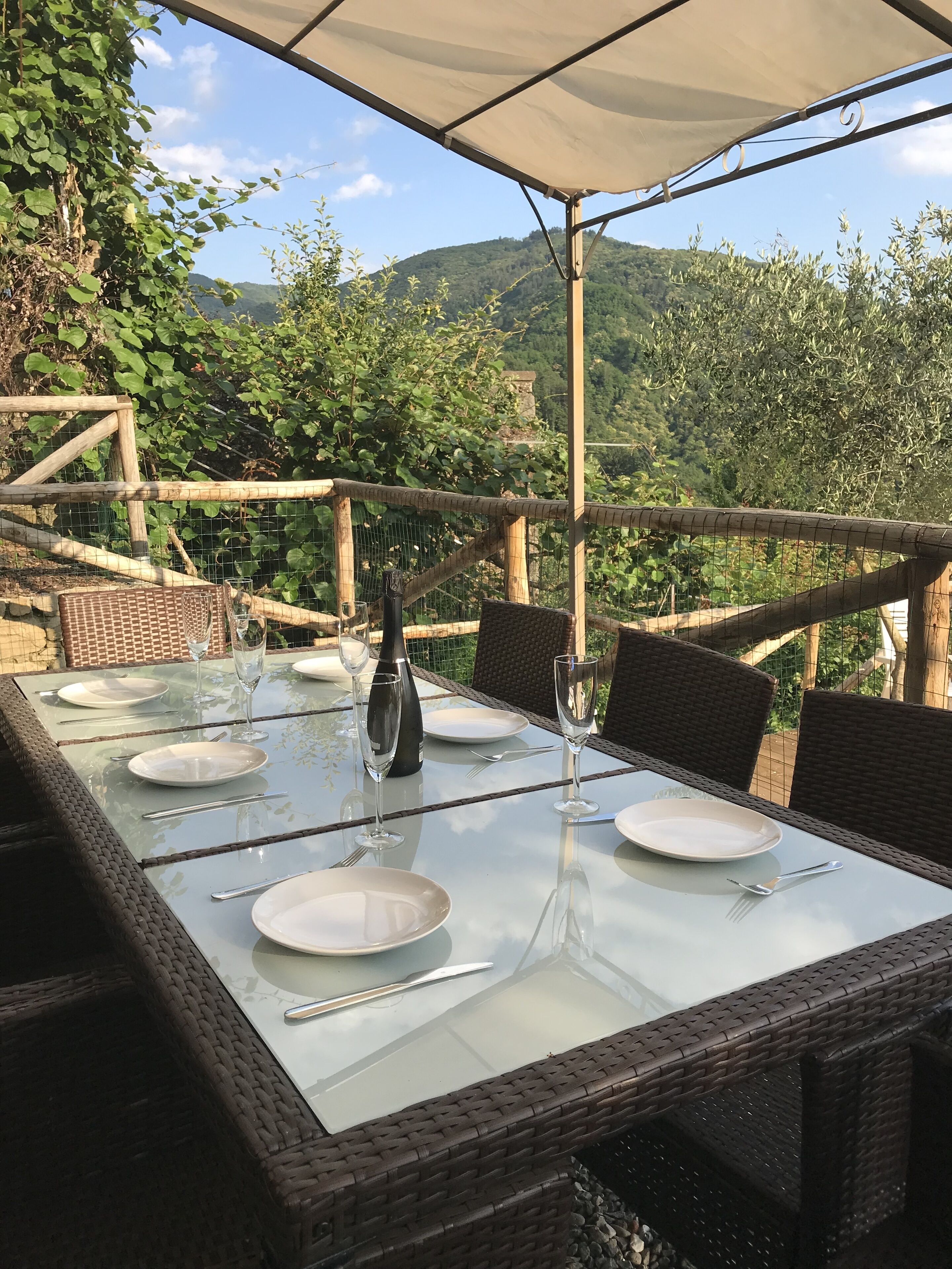 An outdoor dining area with a glass table set for six, surrounded by wicker chairs, overlooking a mountainous landscape.