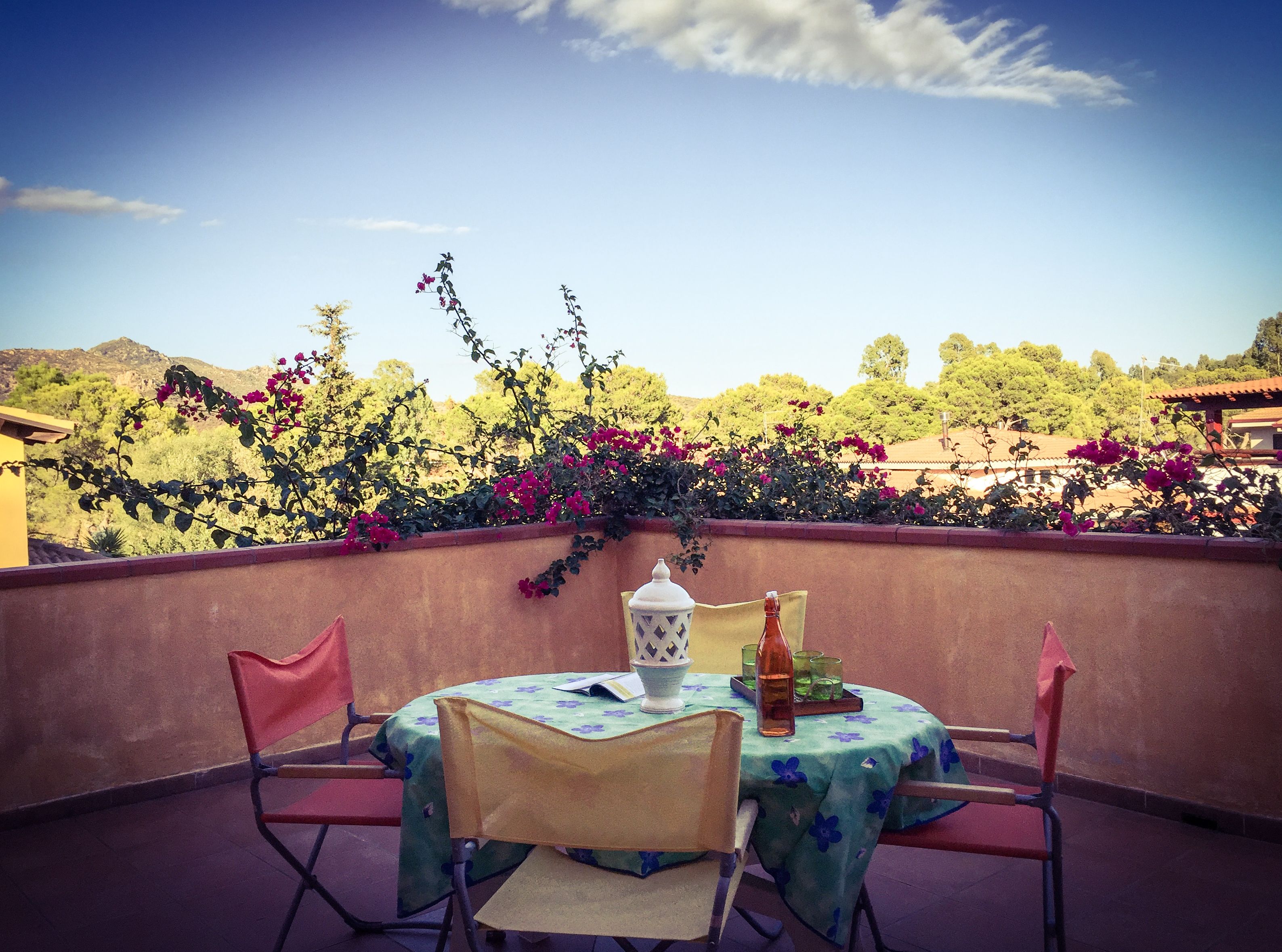 A terrace with a table set for two, surrounded by chairs, overlooking a garden with bougainvillea and a view of a hillside.