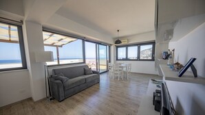 Een ruime woonkamer met een grijze bank, een eettafel met stoelen en grote ramen met uitzicht op zee.