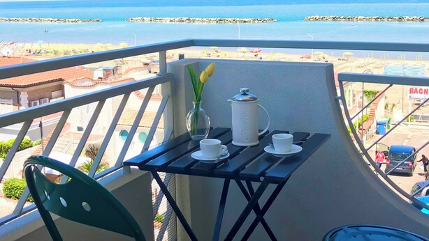Un balcone con un tavolo apparecchiato per due persone, una caffettiera e un vaso con fiori.