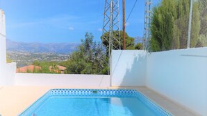 A rectangular swimming pool with a ladder on one side, surrounded by a white wall and a concrete deck. In the background, there are power lines, a tall metal structure, and a view of distant mountains and houses. - Albir Apartment  in House with Community Pool, with the Garden (l'Alfàs del Pi)