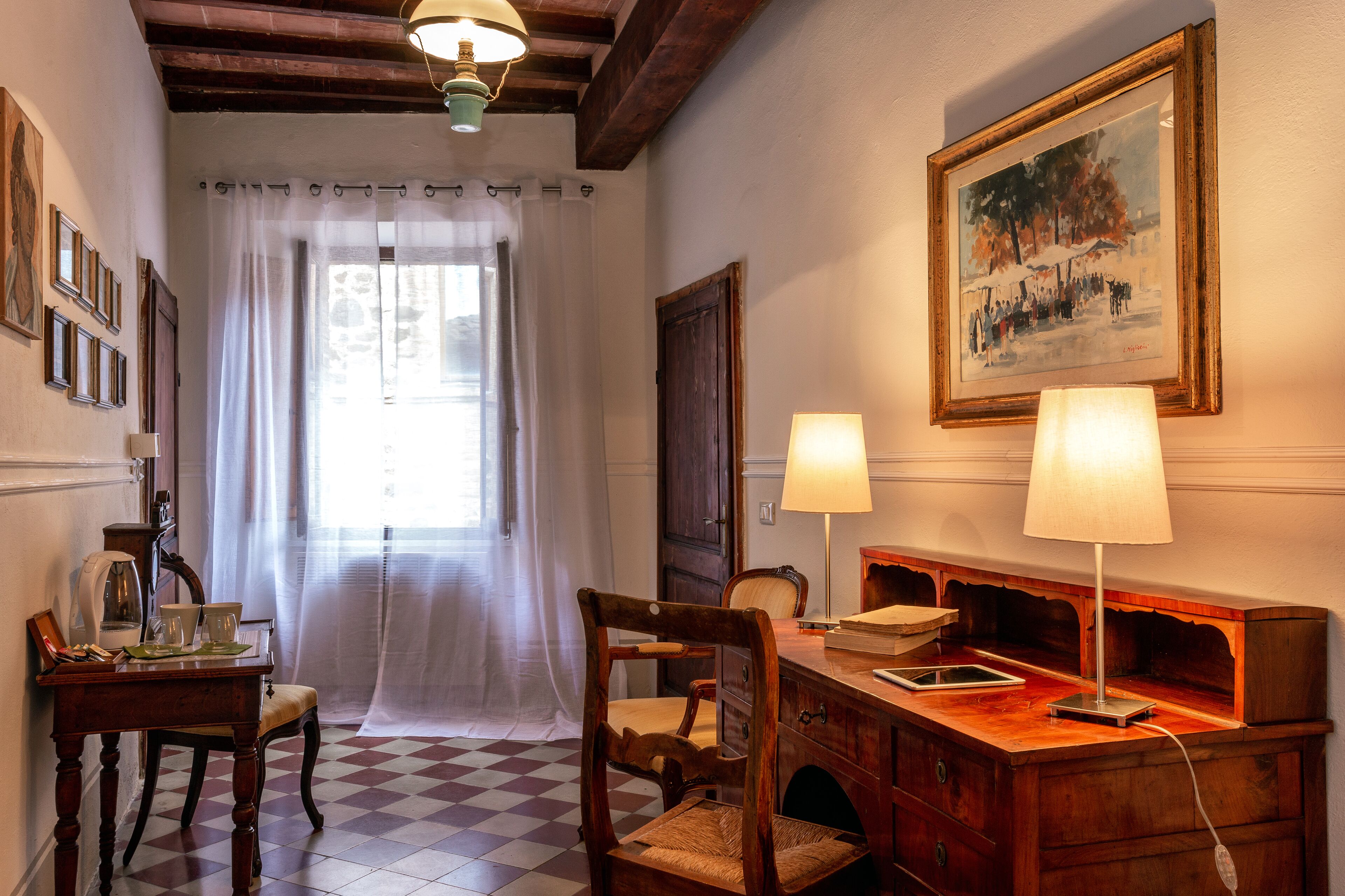 Una habitación con piso a cuadros, un escritorio de madera con lámpara, una silla, una ventana con cortina blanca y un cuadro enmarcado en la pared.
