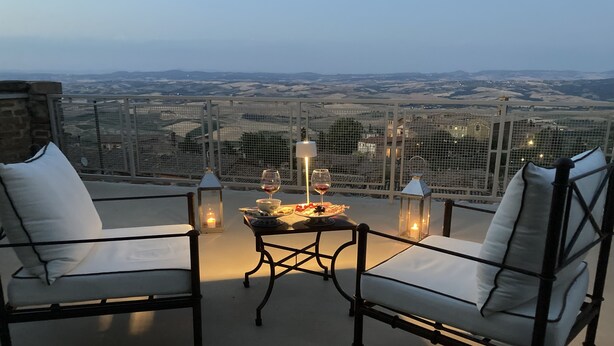 A terrace with two white cushioned chairs, a table with a candle, and a view of distant hills.