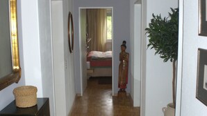 Een gang met een houten vloer, een bed op de achtergrond, een beeld en een spiegel aan de linkerkant.