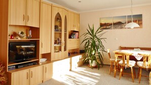 A well-lit room with wooden cabinets, a dining table with chairs, a potted plant, and a painting on the wall.