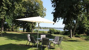 Un espace extérieur aménagé avec une table et des chaises, protégé par un grand parasol, entouré d’arbres et offrant une vue sur un lac.
