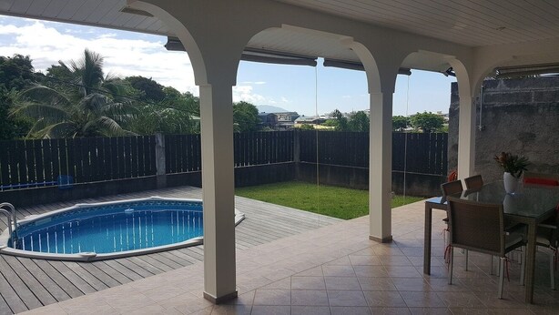 Eine überdachte Terrasse mit Pool, Gartenmöbeln und Blick auf einen umzäunten Garten.