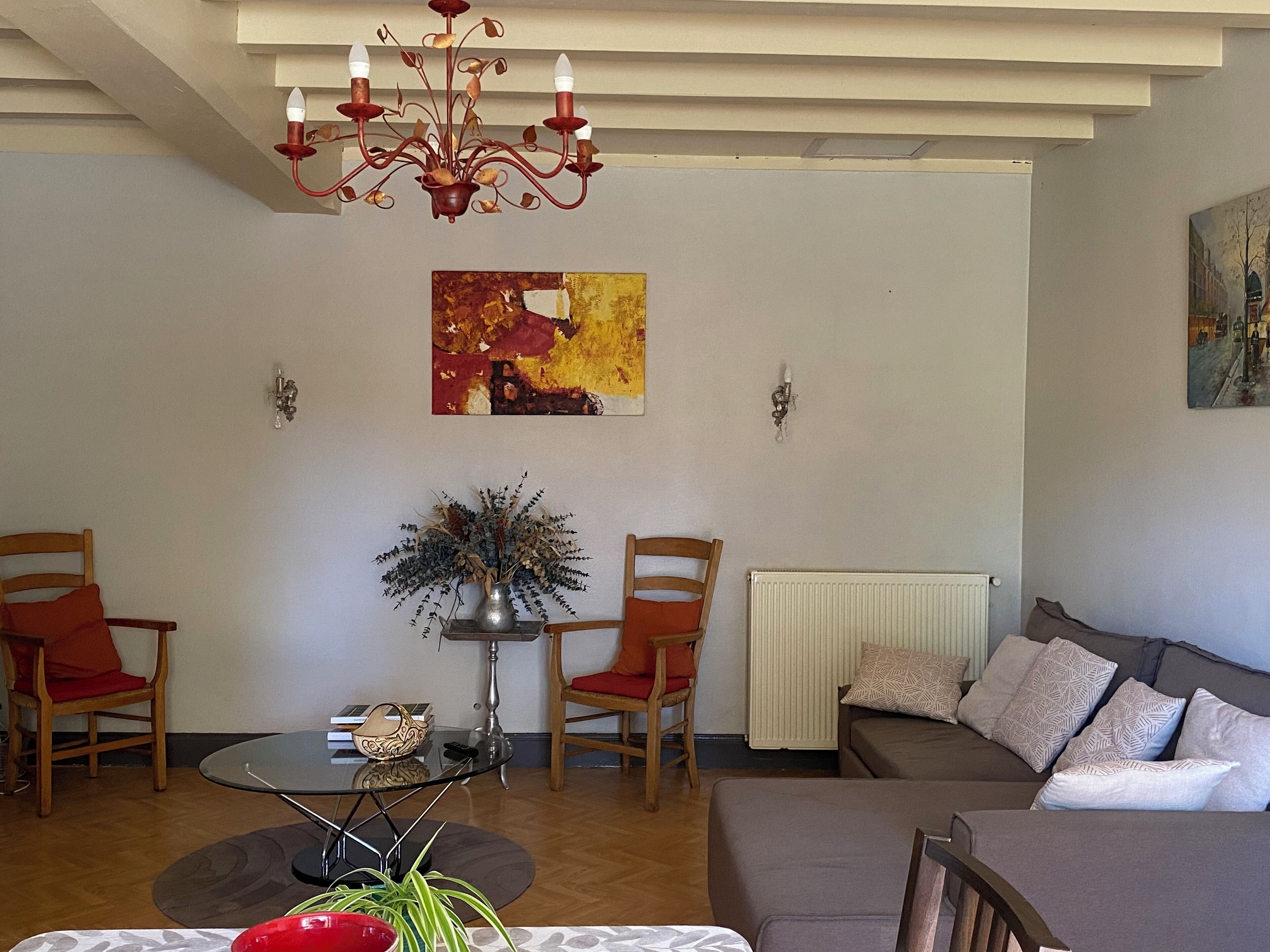 Een woonkamer met een bank, twee stoelen, een tafel met een vaas, een kroonluchter en een schilderij aan de muur.