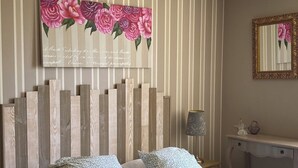 A bedroom with a bed, pillows, a wooden headboard, a wall-mounted mirror, and a decorative floral painting.
