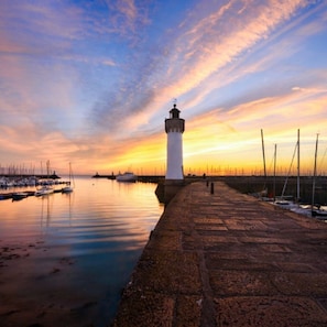 Um farol ao pôr do sol, com águas calmas e barcos ao fundo.