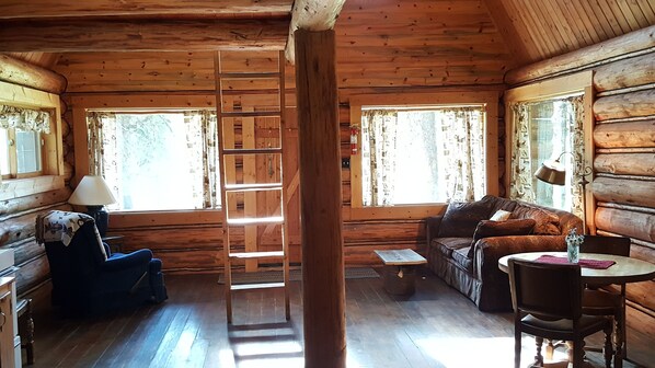 Interior de una cabaña de troncos con piso de madera, un sofá, un sillón, una mesita y una escalera que lleva a un entrepiso.