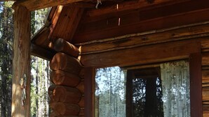 A rustic log cabin with two wicker chairs outside, a wooden porch, and a window with curtains.