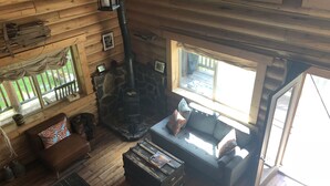 A cozy cabin interior with a stone fireplace, wooden walls, a sofa, and a window overlooking a deck.