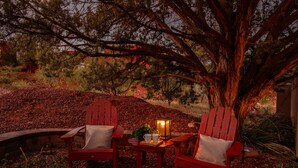 Dos sillones Adirondack rojos con cojines, una mesita con un farol, y un fondo de árboles y follaje.