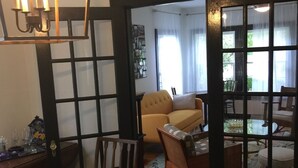 Una zona de menjador amb taula i cadires de fusta, un lampadari i vista cap a un saló amb sofà i cadires a través d'una porta de vidre.